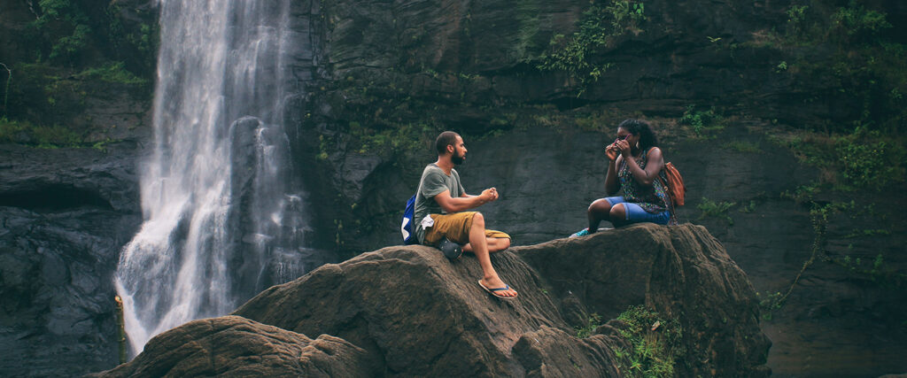 A Black couple in nature, sitting on some boulders next to a waterfall